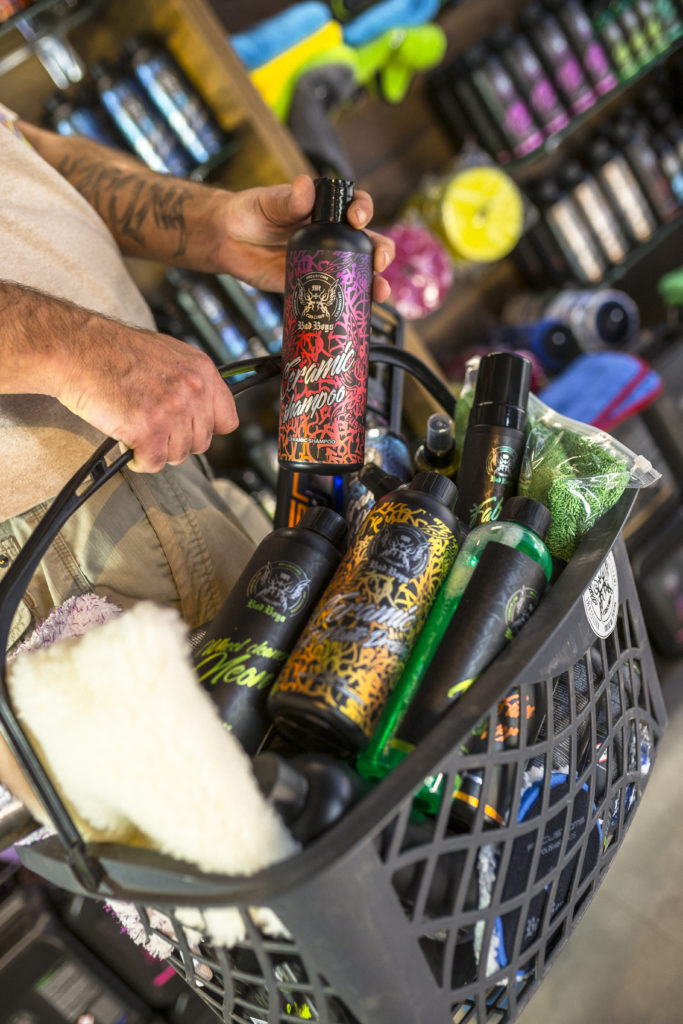 Photo of a customer shopping at RRCustoms. The customer has a shopping cart and a bottle of BadBoys shampoo in his hand. The photo is made on the background of the products standing on the RRCustoms company shelf